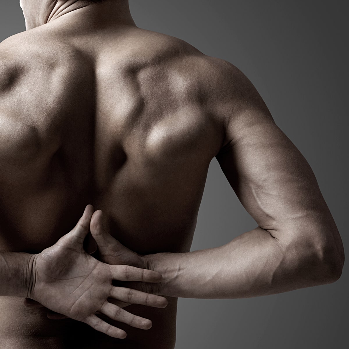 © Ortho Health Institut in München Eine Person ohne Hemd mit muskulösem Rücken und muskulösen Armen wird von hinten gezeigt. Der linke Arm ist hinter dem Rücken, während die rechte Hand den Arm knapp über dem Ellenbogen umfasst. Der Hintergrund, der das Ortho Health Institut in München darstellt, verblasst von Dunkelgrau zu Hellgrau.