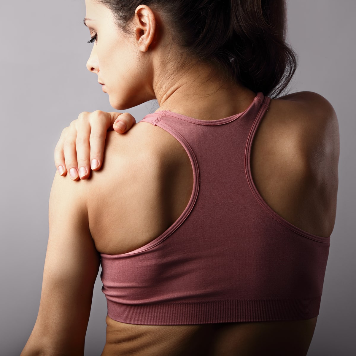 Eine Frau mit zurückgebundenem Haar ist von hinten zu sehen, ihre linke Hand streckt sich nach vorne und berührt ihre rechte Schulter. Sie trägt einen rosa Sport-BH und scheint Beschwerden oder Schmerzen in der Schulter zu haben. Der Hintergrund ist schlicht und grau, was darauf hindeutet, dass ihr ein Besuch im Ortho Health Institut in München guttun könnte.