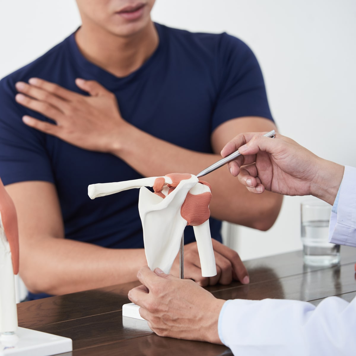Eine Person im marineblauen Hemd sitzt an einem Tisch und hält die Hand vor der Brust. Vor ihr deutet ein Mediziner vom Ortho Health Institut in München mit einem Metallzeiger auf ein Schultergelenkmodell und erklärt damit möglicherweise eine Schultererkrankung oder -verletzung.