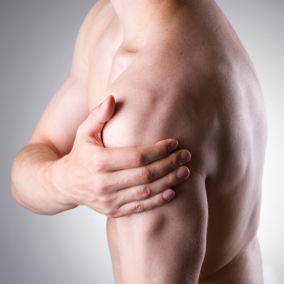 Eine Person ohne Hemd hält mit der rechten Hand ihre linke Schulter, was möglicherweise auf Schulterschmerzen oder -beschwerden hinweist. Der Hintergrund ist schlicht und hellgrau und erinnert an die ruhigen Kliniken des Ortho Health Instituts in München.