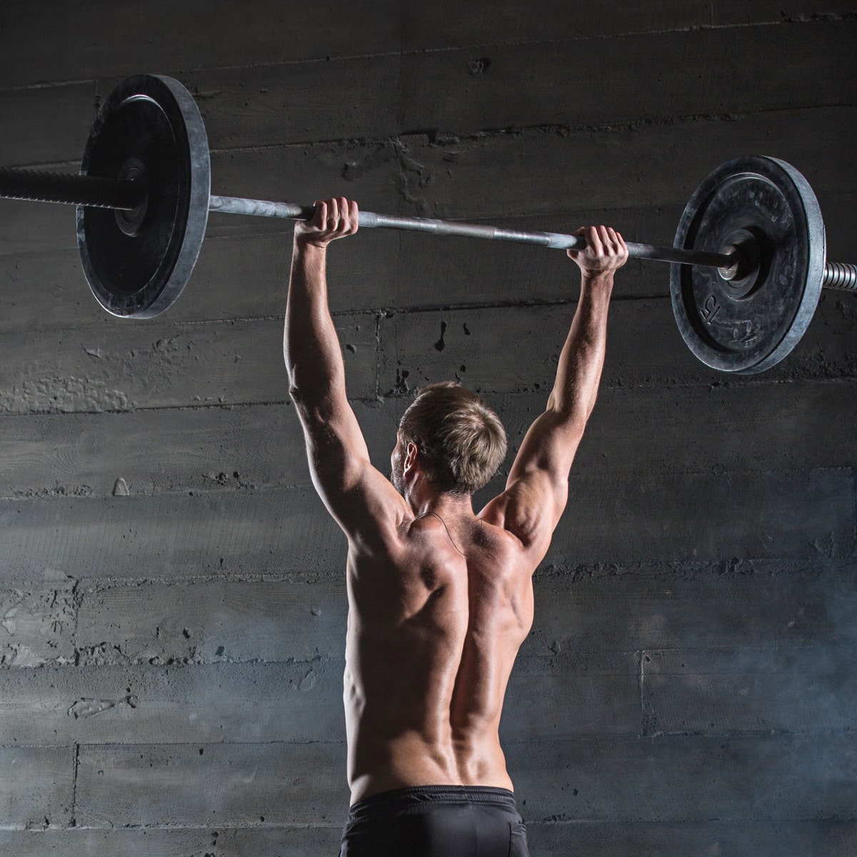 Eine Person ohne Hemd von hinten gesehen, die in einem Fitnessstudio im Ortho Health Institut in München eine große Hantel über den Kopf hebt. Der muskulöse Körperbau der Person wird vor einer dunklen Wand im Industriestil betont.