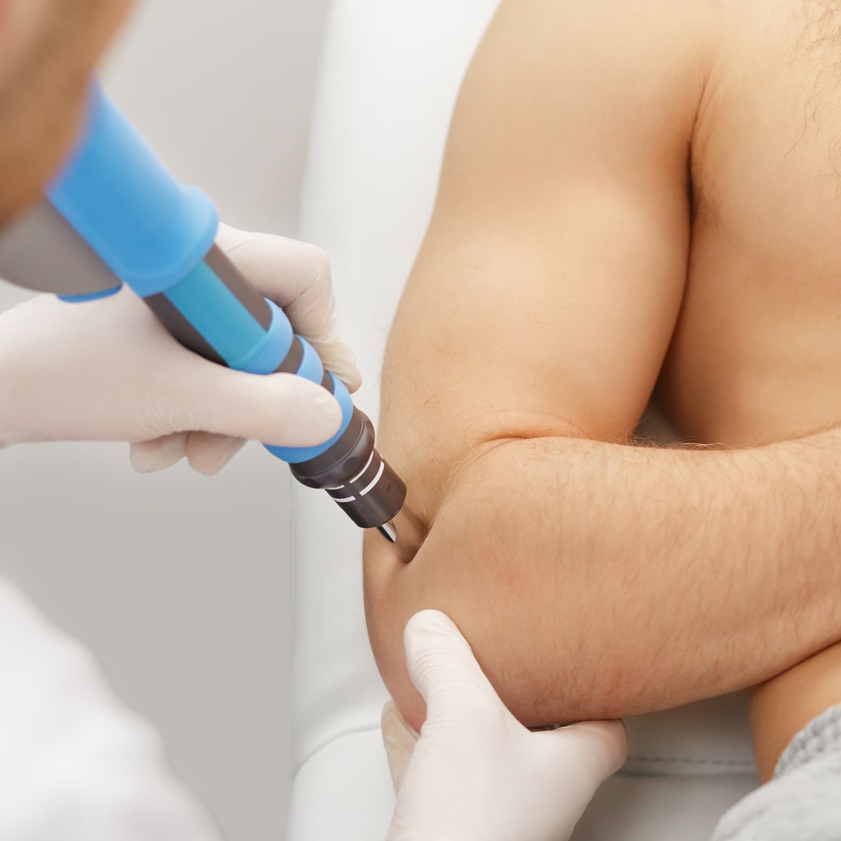 Ein medizinischer Mitarbeiter des Ortho Health Instituts in München verwendet ein blau-graues Gerät, um den Ellenbogen eines Patienten zu behandeln. Der Mitarbeiter, der weiße Handschuhe trägt, richtet das Gerät auf die Innenseite des Ellenbogens des Patienten, während sich der Patient zurücklehnt und den Arm für die Behandlung freigibt.