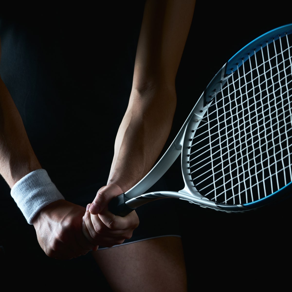 Eine Person mit einem weißen Armband mit der Aufschrift „Ortho Health Institut“ hält einen Tennisschläger mit beiden Händen und hält ihn in einer Position, als ob sie sich auf einen Rückhandschlag vorbereiten würde. Der dunkle Hintergrund hebt das Motiv und den Tennisschläger hervor, während dramatische Beleuchtung den Arm und den Schläger hervorhebt und so eine intensive Szene erzeugt.