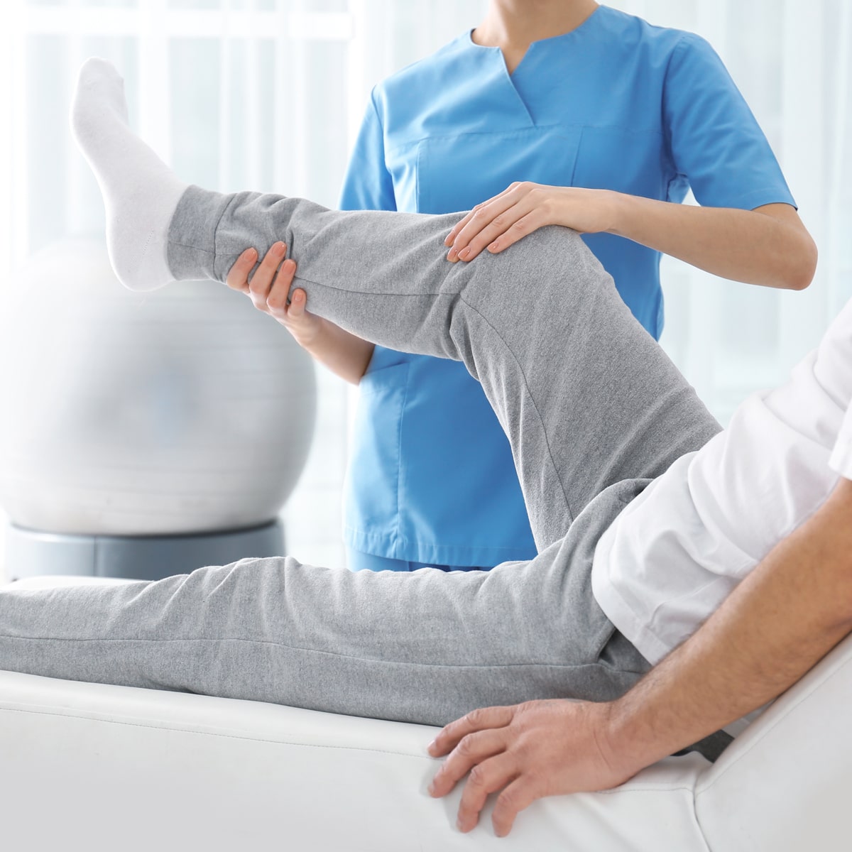 © Ortho Health Institut in München Ein Physiotherapeut in blauer Uniform hilft einem Patienten in grauer Jogginghose und weißen Socken bei einer Beinübung im Ortho Health Institut in München. Der Patient liegt auf einer weißen Liege, während der Therapeut das angewinkelte Bein des Patienten anhebt. Im Hintergrund ist ein großer Gymnastikball zu sehen.