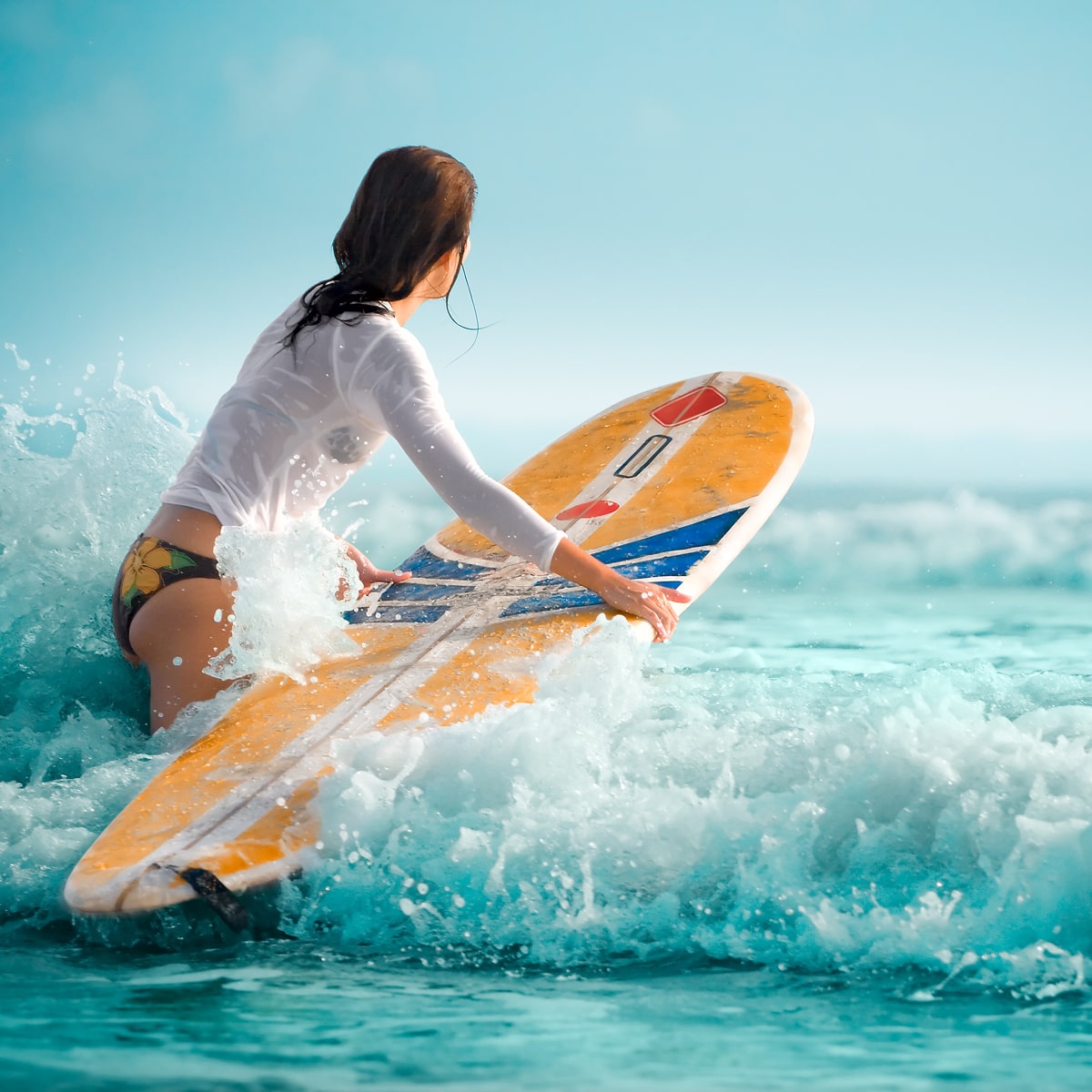 Eine Person mit langen Haaren watet in der Nähe des Ortho Health Instituts München ins Meer und hält ein gelbes Surfbrett in der Hand. Sie trägt ein weißes Oberteil und ein gemustertes Bikinihöschen. Das Surfbrett ist teilweise unter Wasser, während kleine Wellen um sie herum plätschern, während Himmel und Meer einen leuchtenden Türkiston aufweisen.