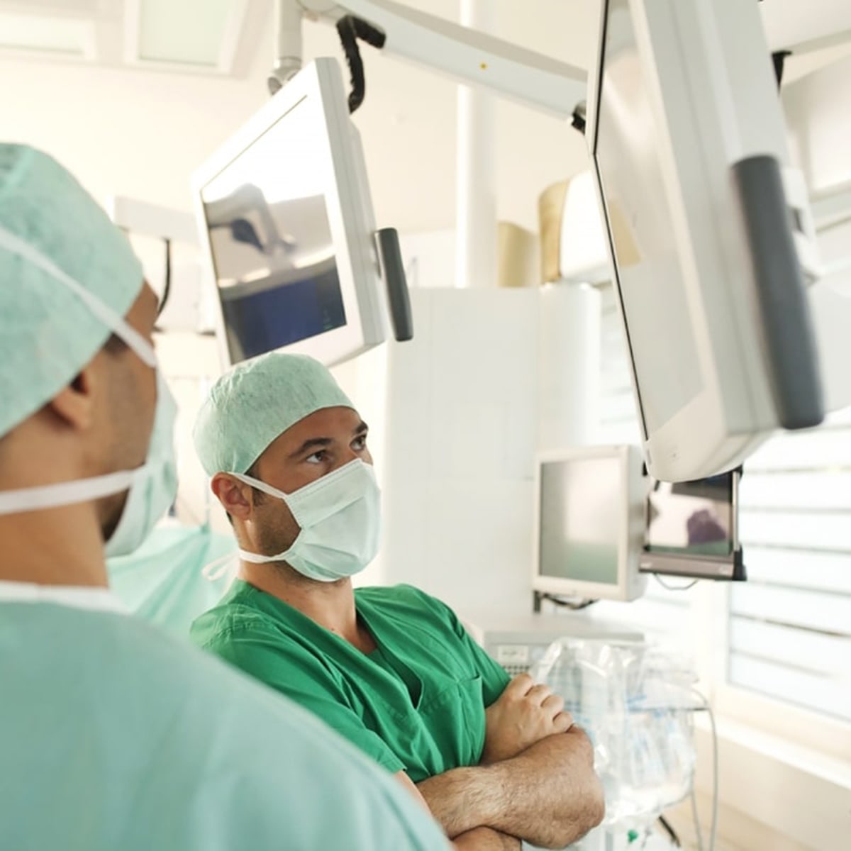 Zwei Chirurgen in grünen OP-Kitteln und mit Gesichtsmasken arbeiten am Ortho Health Institut in München in einem Operationssaal und konzentrieren sich auf Monitore, die von der Decke hängen. Ein Chirurg hat die Arme verschränkt, während im Hintergrund verschiedene medizinische Geräte und Maschinen zu sehen sind.