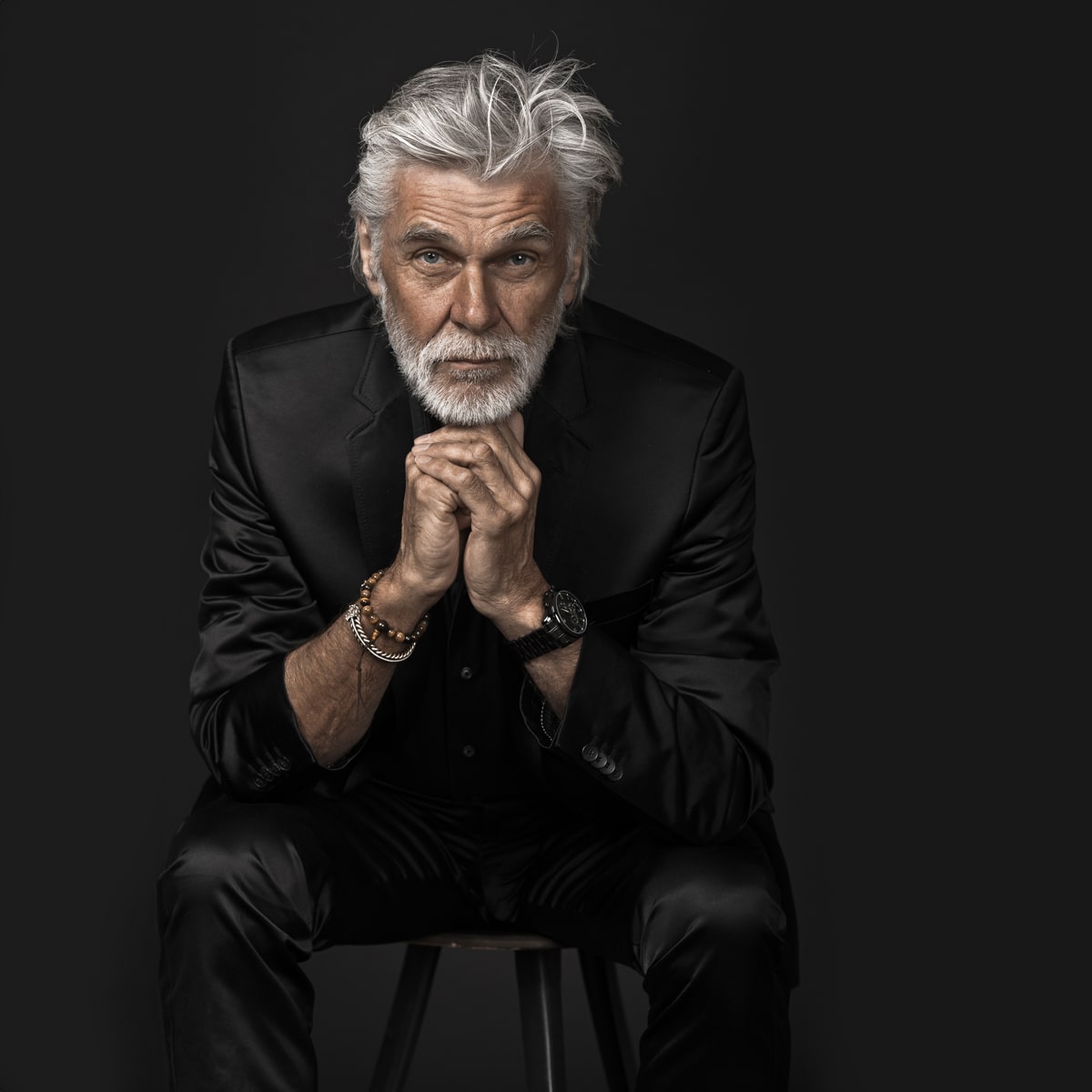 © Ortho Health Institut in München Ein vornehmer älterer Herr mit silbernem Haar und Bart, gekleidet in einen schwarzen Satinanzug, sitzt auf einem Hocker vor einem dunklen Hintergrund. Er stützt sein Kinn auf seine Hände, die mit Armbändern und einer Uhr aus München geschmückt sind, und blickt intensiv in die Kamera.