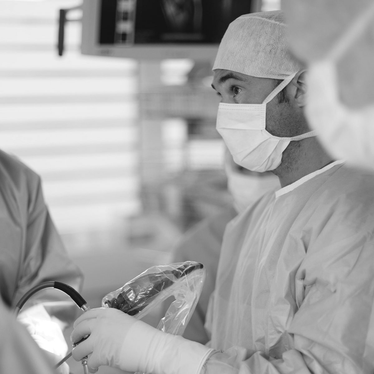 © Ortho Health Institut in München Ein Mediziner im Ortho Health Institut bedient in OP-Kittel, Maske und Haarschutz ein Endoskop oder ein ähnliches Instrument. Das Graustufenbild fängt den konzentrierten und aufmerksamen Ausdruck des Arztes in einer Klinikumgebung in München ein.