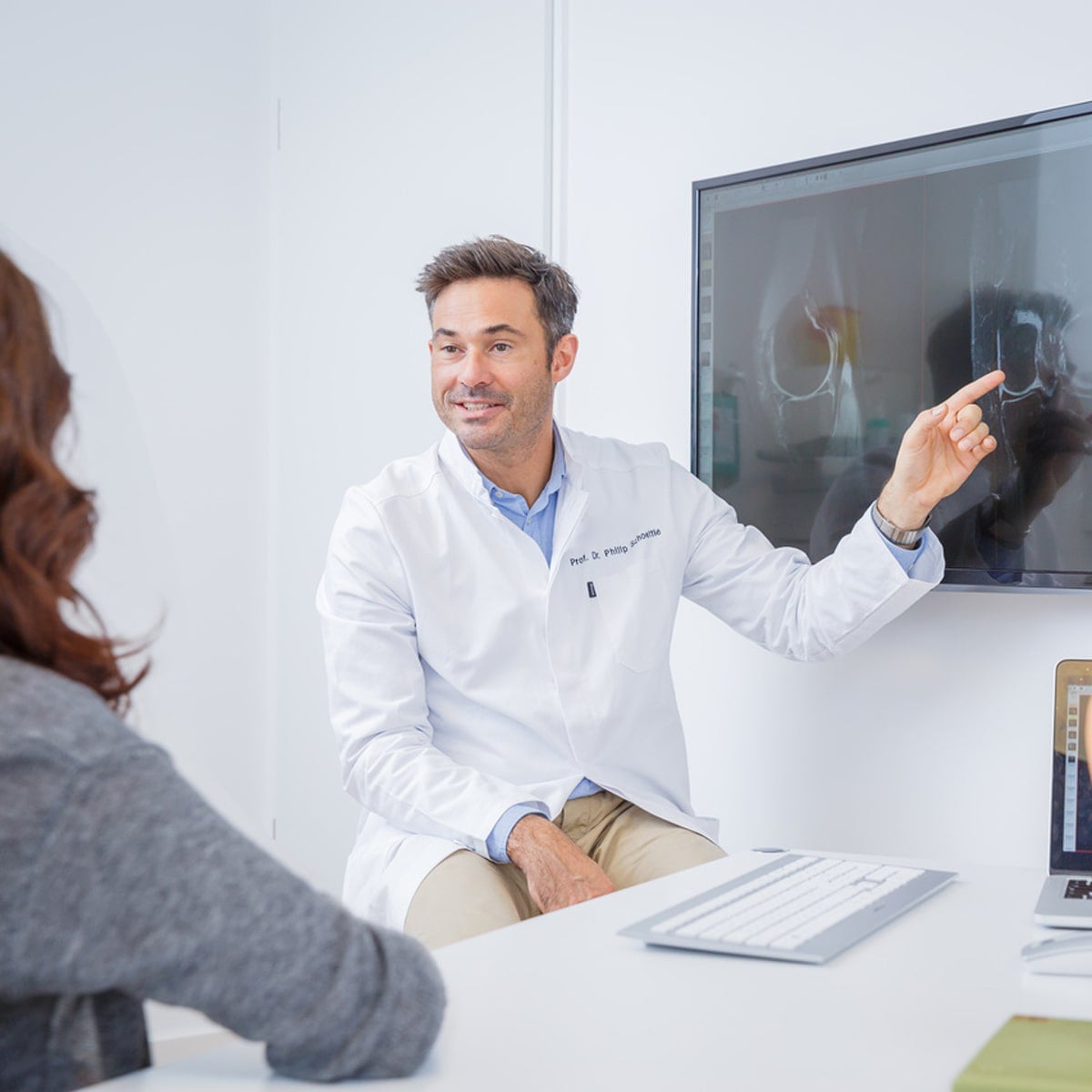 © Ortho Health Institut in München Ein Arzt im weißen Kittel sitzt an einem Schreibtisch im Ortho Health Institut in München und zeigt auf ein Röntgenbild, das auf einem Monitor angezeigt wird. Er hat einen freundlichen Gesichtsausdruck und erklärt einer ihm gegenübersitzenden Patientin etwas. Auf dem Schreibtisch liegen eine Tastatur und ein Laptop.