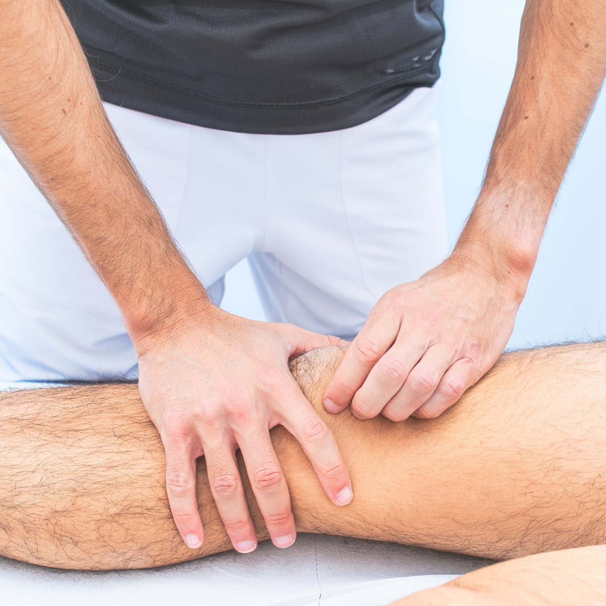 © Ortho Health Institut in München Ein Physiotherapeut vom Ortho Health Institut in München, der ein schwarzes Hemd und weiße Hosen trägt, behandelt das Bein eines Patienten. Die Hände des Therapeuten konzentrieren sich auf das Knie des Patienten und üben Druck auf den Bereich aus, während der Patient auf einem Behandlungstisch liegt.