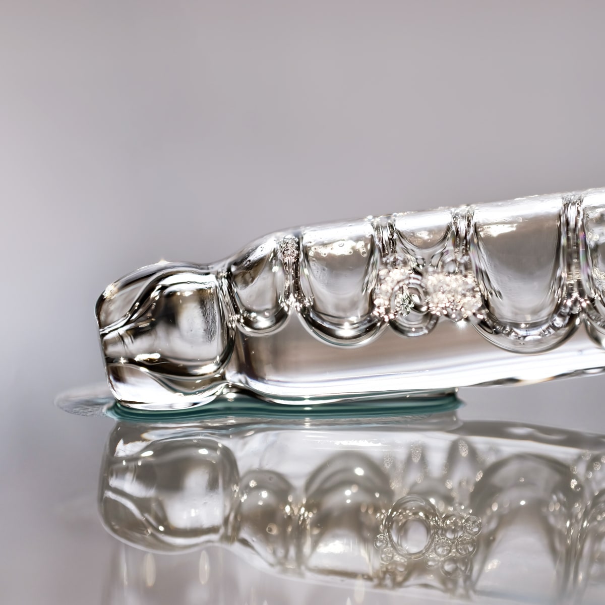 © Ortho Health Institut in München Nahaufnahme eines klaren Glasobjekts, das auf einer reflektierenden Oberfläche liegt. Das Objekt hat ein kompliziertes Design mit wellenförmigen Mustern und Blasen, die ihm ein zartes und kompliziertes Aussehen verleihen. Die Reflexion auf der Oberfläche hebt seine detaillierte Handwerkskunst hervor und erinnert an die feine Kunstfertigkeit, die in München zu finden ist.