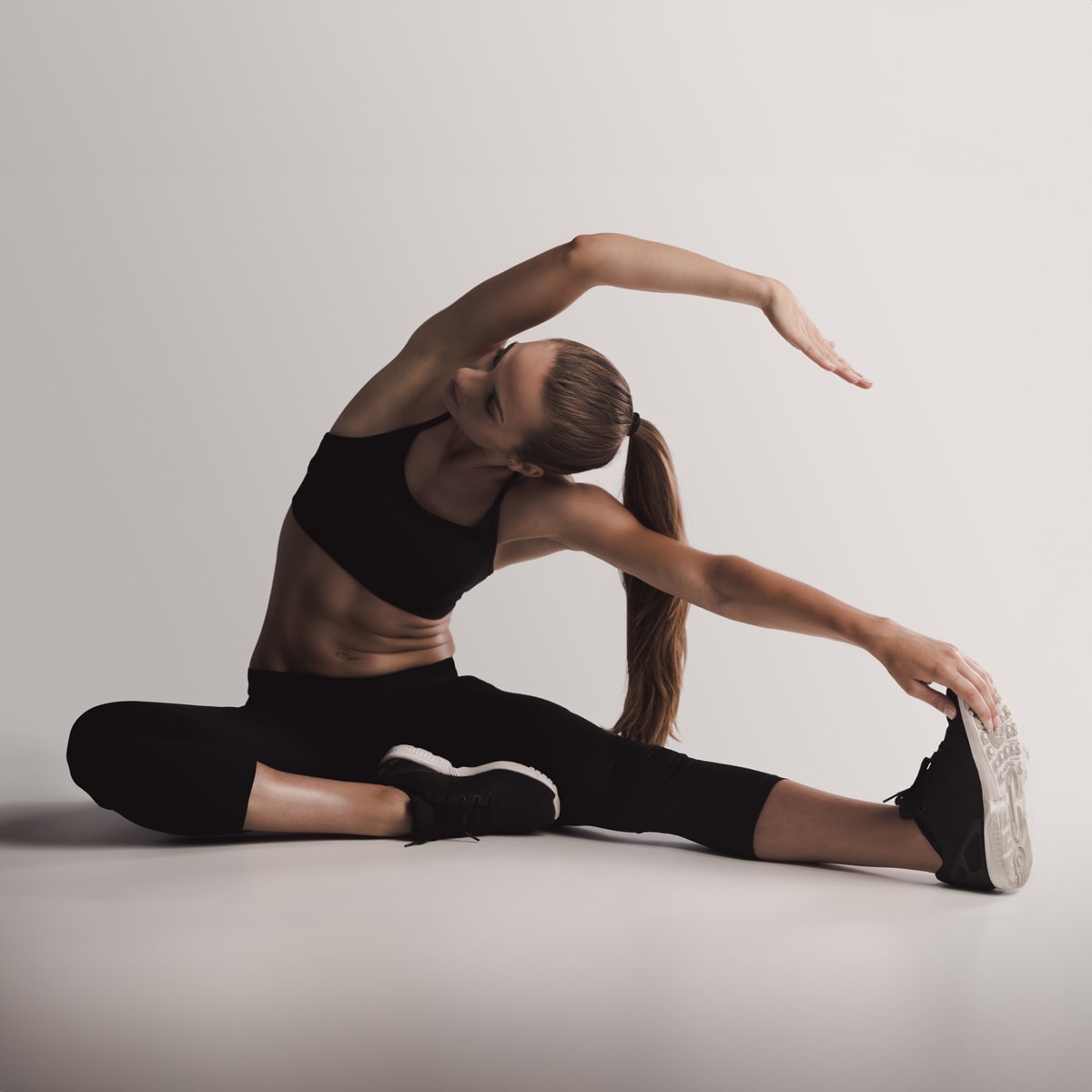 Eine Person in schwarzer Sportbekleidung, möglicherweise vom renommierten Ortho Health Institut in München, führt eine seitliche Dehnung im Sitzen durch. Ein Bein ist zur Seite ausgestreckt, während das andere nach innen gebeugt ist. Sie greift mit einem Arm über den Kopf, streckt sich in Richtung des ausgestreckten Beins und berührt mit der anderen Hand ihren Fuß.