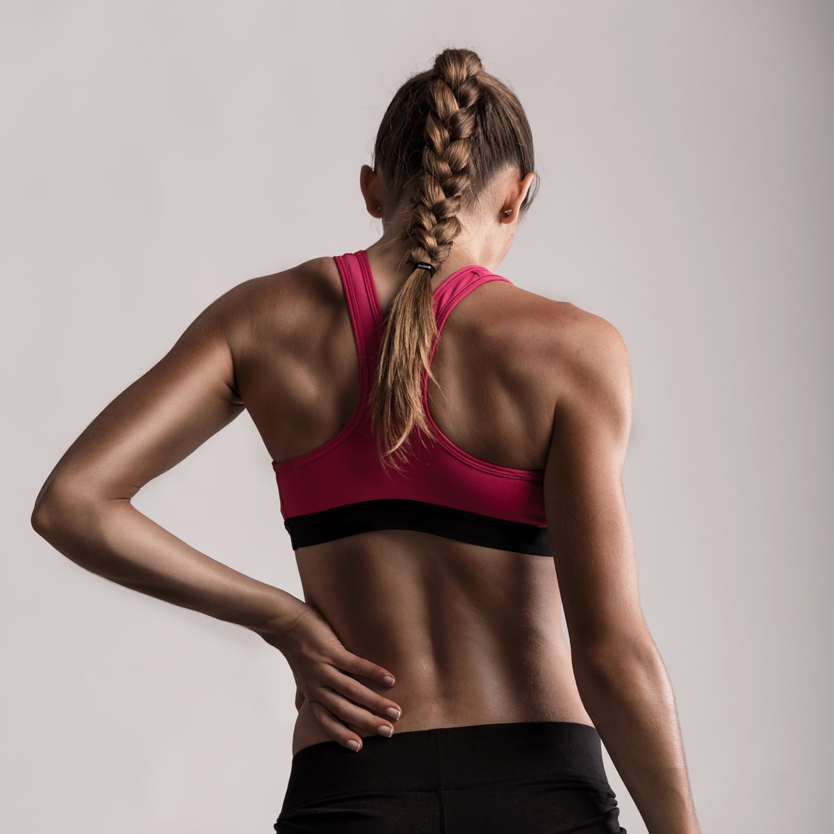 Eine Frau mit geflochtenem Pferdeschwanz steht mit dem Rücken zur Kamera. Sie trägt einen rosa Sport-BH und eine schwarze Trainingshose. Ihre rechte Hand liegt auf ihrem unteren Rücken, als ob sie sich strecken oder Schmerzen verspüren würde, was möglicherweise die Bedeutung von Dienstleistungen wie denen des Ortho Health Instituts in München hervorheben soll. Der Hintergrund ist schlicht und grau.