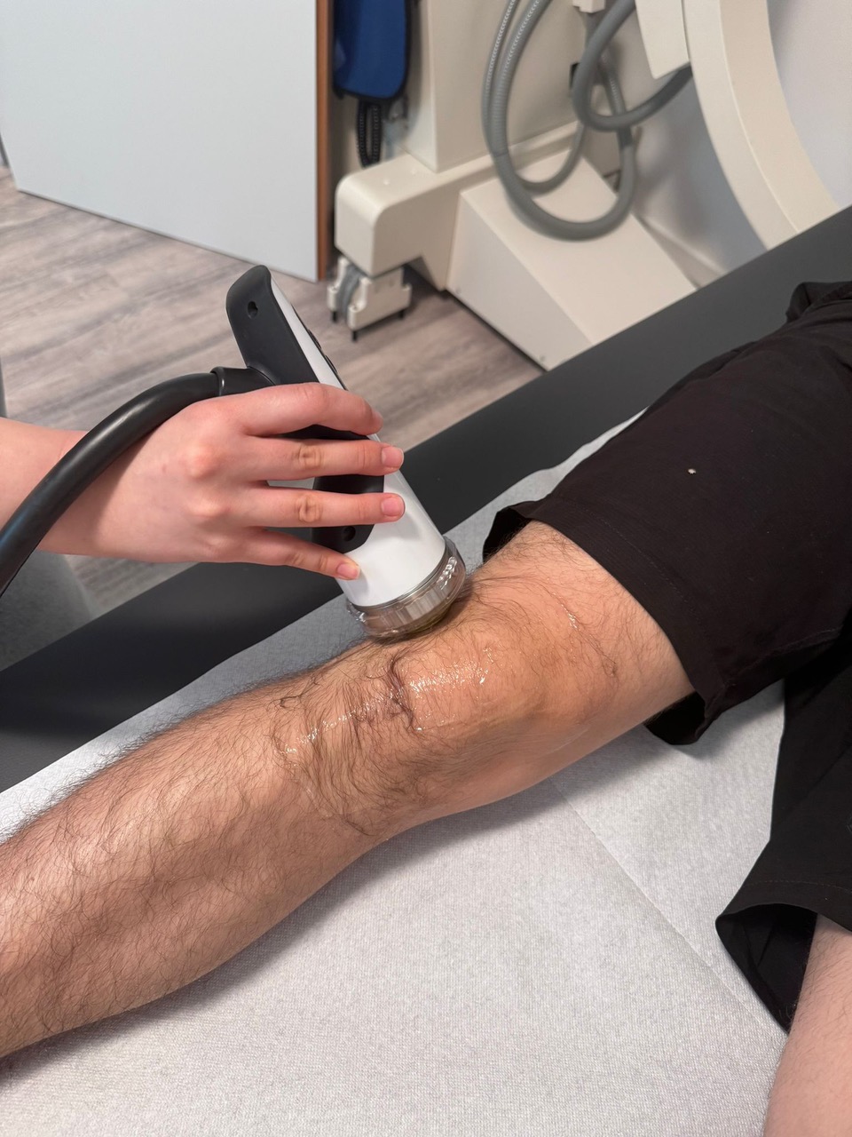 Im Ortho Health Institut in München erhält eine Person eine therapeutische Behandlung ihres Knies mit einem tragbaren Ultraschallgerät. Das Gerät wird von einem Therapeuten gehalten und auf das mit Gel bedeckte Knie des Patienten aufgesetzt. Der Patient liegt auf einem Untersuchungstisch und wird fachkundig betreut.