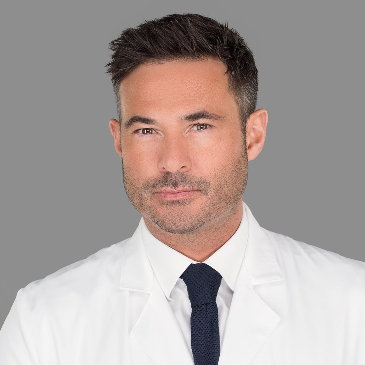 Ein Mann mit kurzen, dunklen Haaren und hellem Bartstoppeln blickt in die Kamera. Er trägt einen weißen Arztkittel über einem weißen Oberhemd mit dunkler Krawatte. Der Hintergrund ist in einem satten Grau gehalten, was auf seinen beruflichen Arbeitsplatz am Ortho Health Institut in München hinweist.
