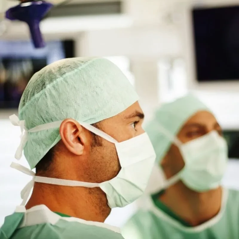 Ortho-Health-Institut-Muenchen-Privatpraxis-Kniespezialist-Sportmedizin-Kompetenz-Patella-Schoettle-Point-Gespiegelt-800×800 Zwei Chirurgen des Ortho Health Instituts sind in einem Operationssaal in München zu sehen. Sie tragen grüne OP-Kittel, Hauben und Masken. Ein Chirurg steht im Vordergrund, während der andere im Hintergrund leicht verschwommen ist. Im unscharfen Hintergrund sind medizinische Geräte und Monitore zu sehen.
